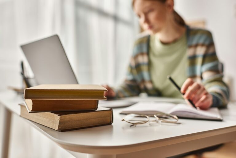 focus on books on desk, teenage girl holding pen and looking at laptop while studying from home